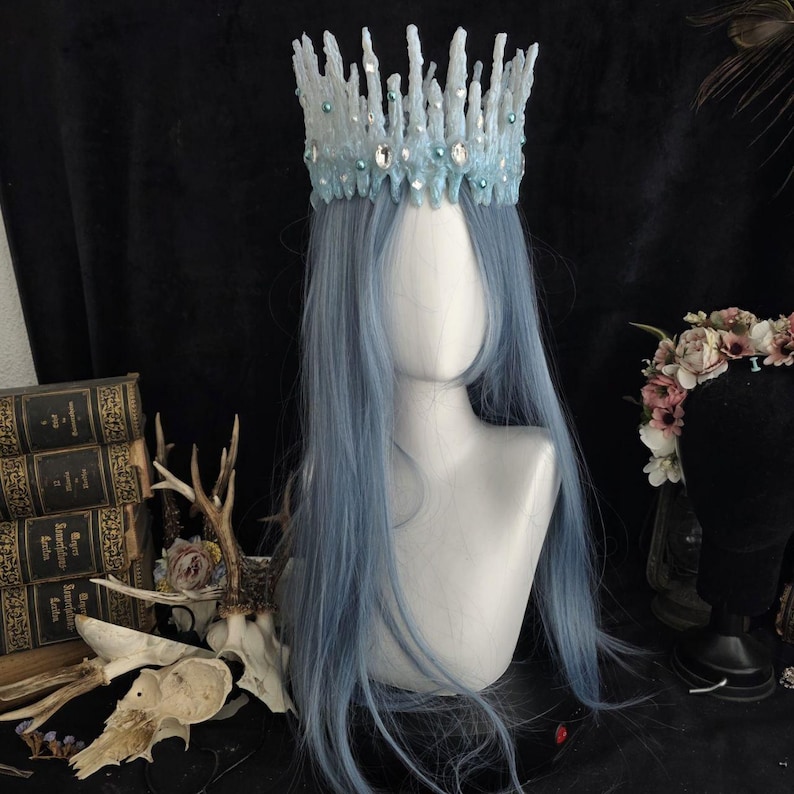 May include: A light blue crown with a jagged, ice-like texture, adorned with clear crystals and small teal beads. The crown is displayed on a mannequin head featuring long, straight, light blue hair, creating a striking and detailed headpiece.