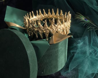 Puede incluir: Una corona dorada con una superficie texturizada, irregular y orgánica que presenta puntas afiladas. La corona está colocada dentro de una caja redonda de terciopelo verde oscuro con tapa, acentuada con tela verde y una pluma de pavo real.