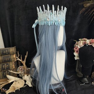 May include: A light blue crown featuring a jagged, ice-like texture adorned with small white pearls and clear oval crystals. The crown is positioned on a mannequin head with long, straight, light blue hair and includes a matching light blue ribbon.