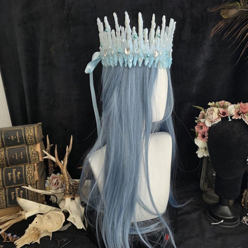 May include: A light blue crown featuring a jagged, ice-like texture adorned with small white pearls and clear oval crystals. The crown is positioned on a mannequin head with long, straight, light blue hair and includes a matching light blue ribbon.