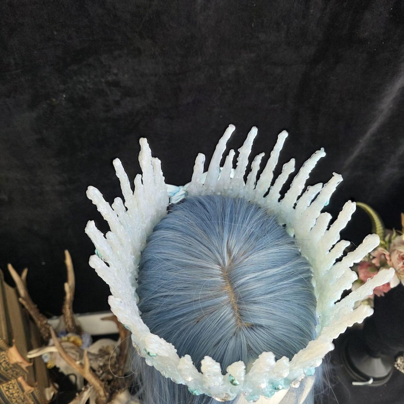 May include: A crown featuring white, jagged, crystal-like spikes arranged in a circular formation. The crown is positioned atop light blue hair, with small, clear, reflective accents embedded along the base of the white, textured structure.