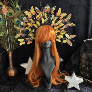 May include: A black headband with a halo of yellow and orange butterflies. The butterflies are made of paper and have a 3D effect. The headband is worn on a mannequin with long, orange hair.