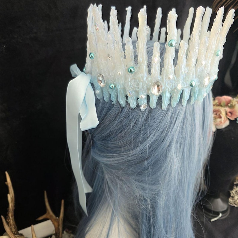 May include: A crown featuring tall, jagged, white and light blue icicle-like spikes. The base is adorned with clear faceted crystals and small, round, light blue beads. A light blue satin ribbon is tied to the side of the crown structure.
