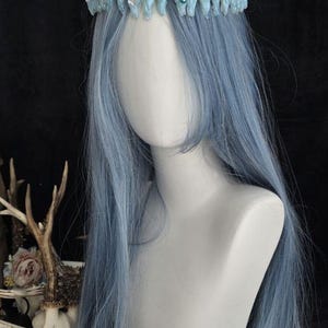 May include: A light blue crown featuring a textured, jagged icicle-like design adorned with small round beads and clear faceted crystals. The crown is positioned on a white mannequin head with long, straight, light blue hair.
