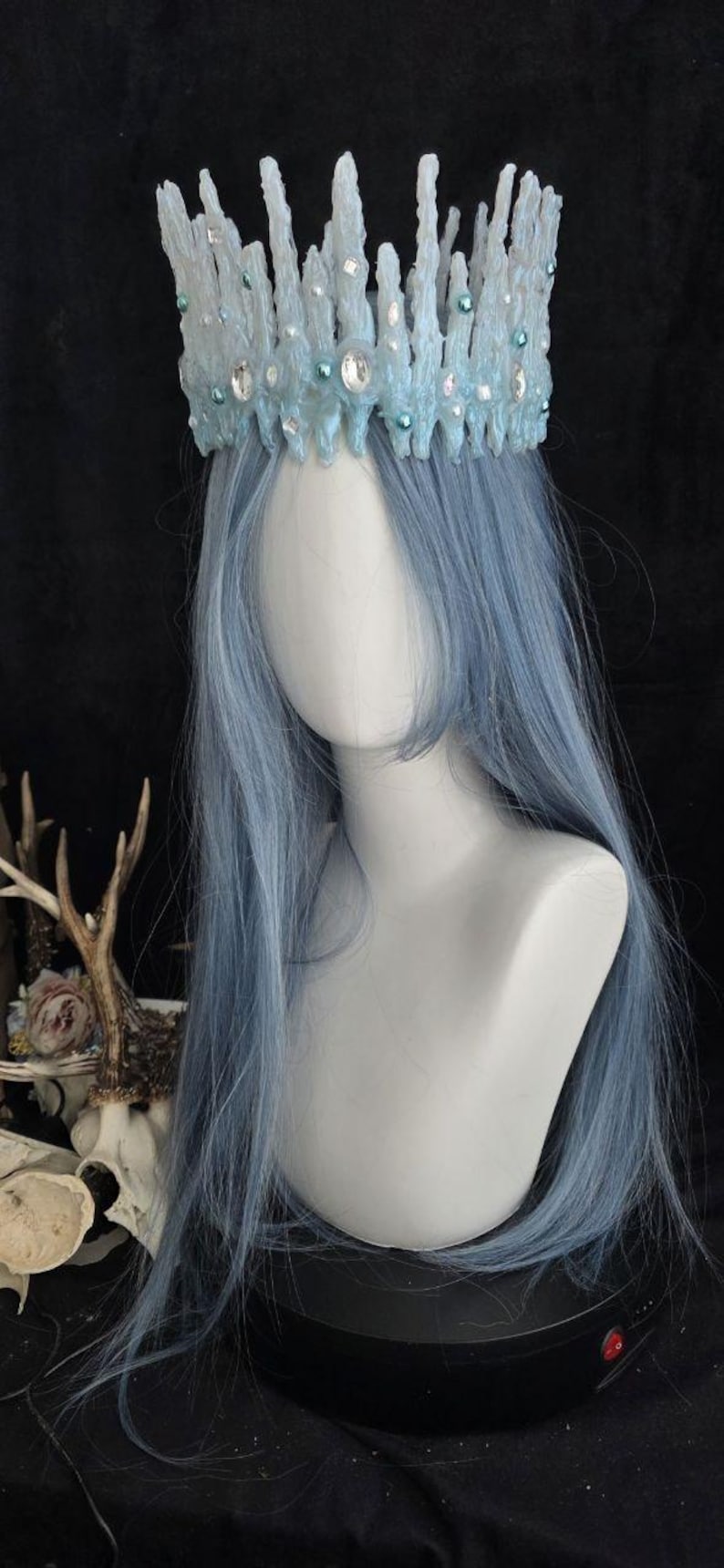 May include: A light blue crown featuring a textured, jagged icicle-like design adorned with small round beads and clear faceted crystals. The crown is positioned on a white mannequin head with long, straight, light blue hair.