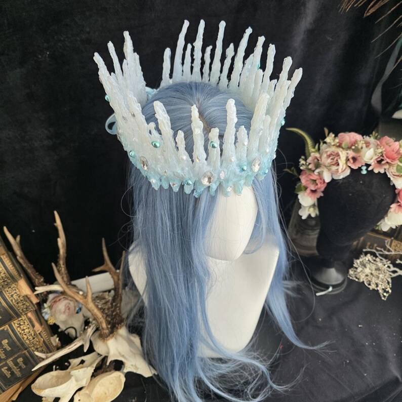 May include: A crown featuring tall, white, icicle-like spikes arranged in a circular formation. The base is adorned with light blue beads and clear, faceted crystals. The crown is displayed on a mannequin head with long, straight, light blue hair.