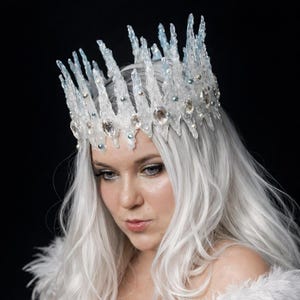 May include: A crown featuring tall, jagged, translucent white and light blue spikes resembling icicles. The base is adorned with clear, faceted crystals and small, round, iridescent beads, creating a textured, crystalline appearance.