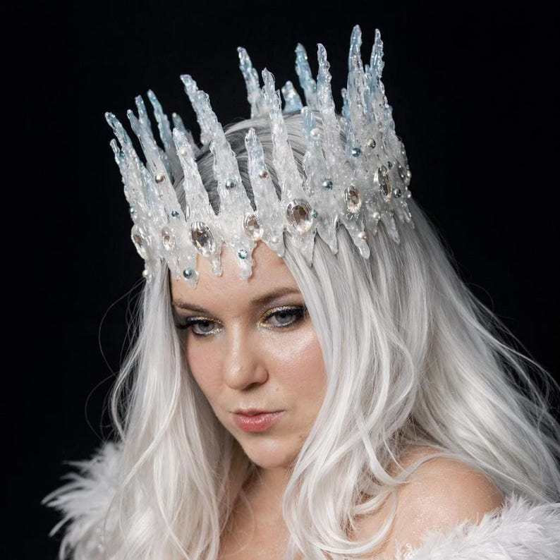 May include: A crown featuring tall, jagged, translucent white and light blue spikes resembling icicles. The base is adorned with clear, faceted crystals and small, round, iridescent beads, creating a textured, crystalline appearance.
