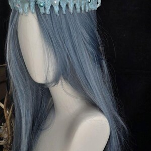 May include: A pale blue crown with a textured, icicle-like structure featuring embedded clear crystals and small teal beads. The crown is positioned atop a mannequin head with long, straight, light blue hair against a dark background.
