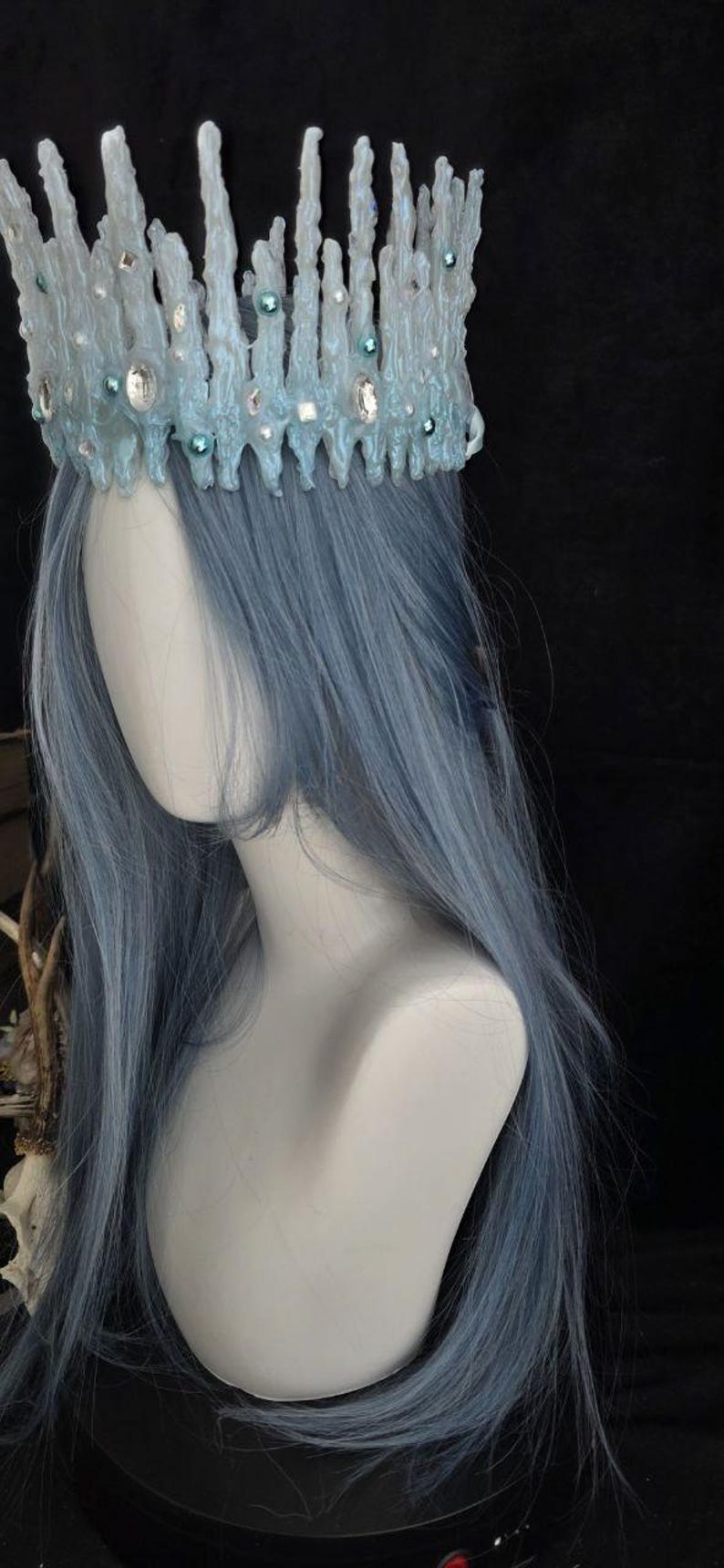 May include: A pale blue crown with a textured, icicle-like structure featuring embedded clear crystals and small teal beads. The crown is positioned atop a mannequin head with long, straight, light blue hair against a dark background.