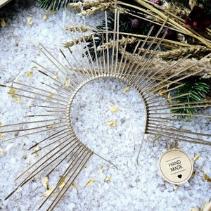 Pode incluir: Uma coroa de halo com design de raios solares em tom dourado, composta por uma tiara circular com inúmeras pontas metálicas radiantes de comprimentos variados. Uma pequena etiqueta redonda bege com "HAND MADE" e um coração está presente.