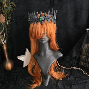 May include: A black crown with green gems on a mannequin head with long, orange hair.