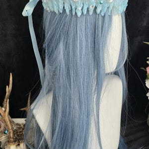 May include: A crown featuring a jagged, icicle-like structure in shades of white and light blue. The crown is adorned with clear faceted crystals, small white pearls, and round teal beads. A light blue ribbon tie hangs from the side of the crown.