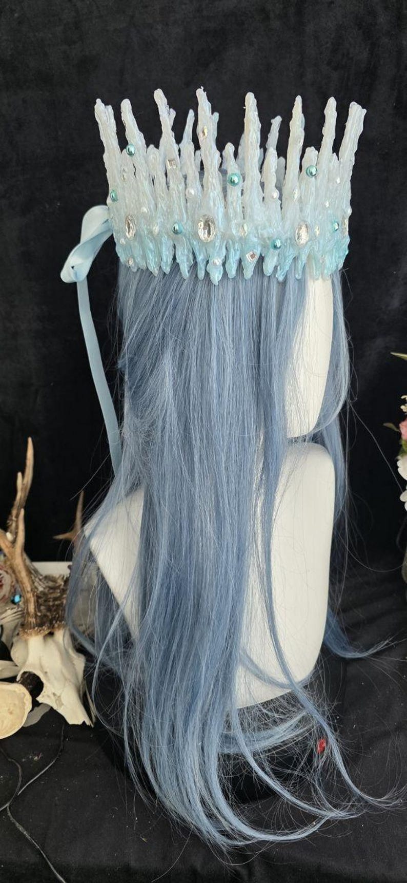 May include: A crown featuring a jagged, icicle-like structure in shades of white and light blue. The crown is adorned with clear faceted crystals, small white pearls, and round teal beads. A light blue ribbon tie hangs from the side of the crown.
