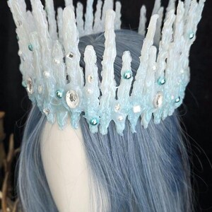 May include: A crown featuring tall, jagged, translucent white and pale blue spikes resembling icicles. The structure is adorned with small, round, light blue beads and clear, faceted crystals embedded throughout the textured, icy surface.