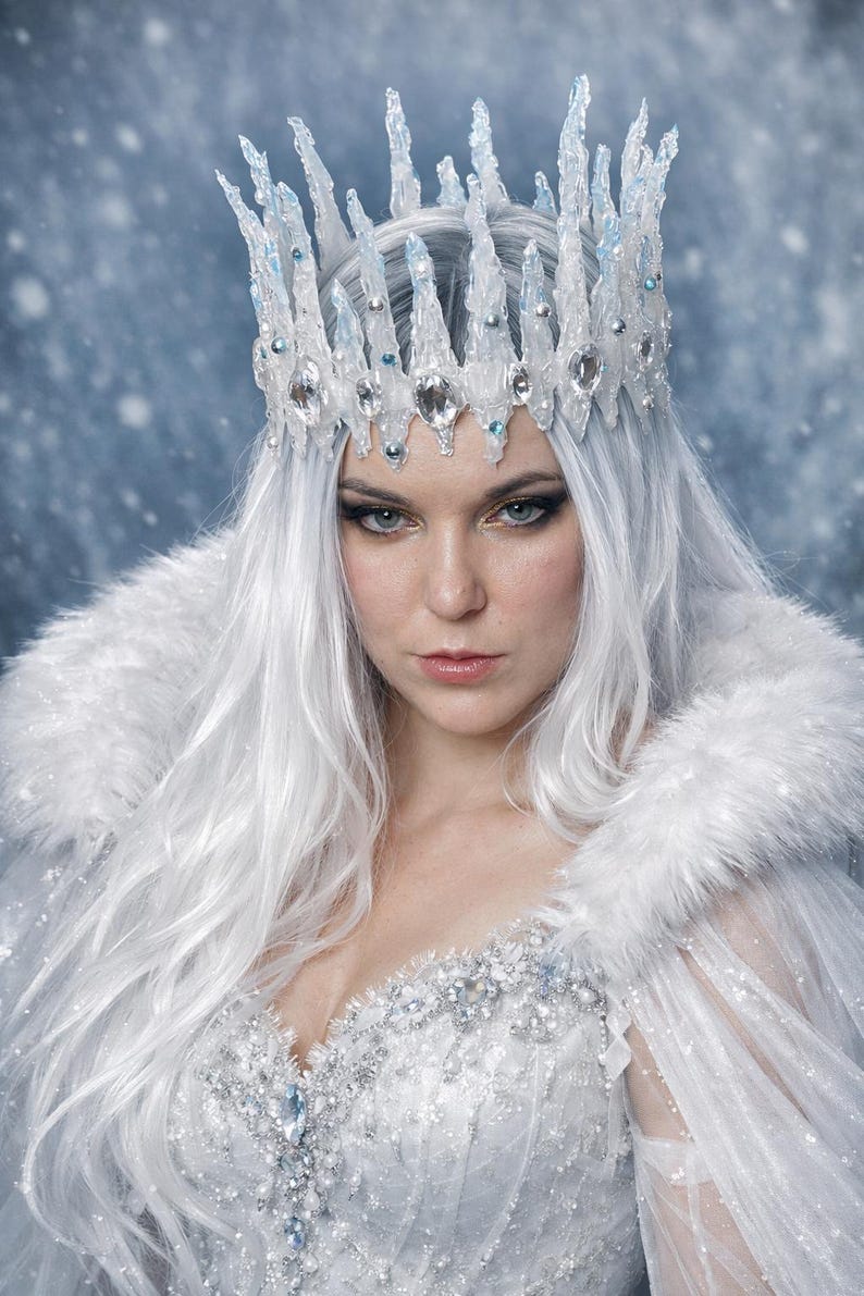 May include: A crown featuring tall, jagged, translucent icicle-like spikes adorned with clear faceted crystals and small silver beads. The crown is worn with a white faux fur-trimmed cape and a bodice embellished with intricate silver beadwork.