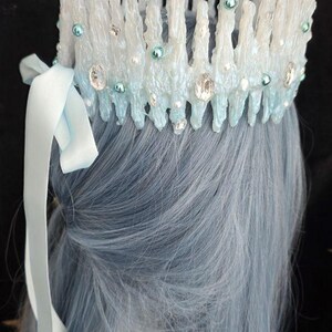 May include: A crown featuring vertical, textured white and light blue spikes resembling icicles. The piece is adorned with small round teal beads, clear faceted crystals, and tiny white pearls. A light blue satin ribbon is tied to the side.