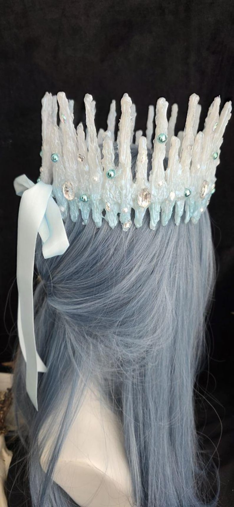 May include: A crown featuring vertical, textured white and light blue spikes resembling icicles. The piece is adorned with small round teal beads, clear faceted crystals, and tiny white pearls. A light blue satin ribbon is tied to the side.