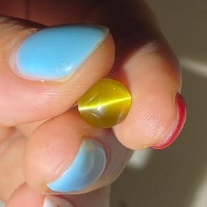以下が含まれることがあります： 猫目効果のある丸い黄色の宝石。青いマニキュアと赤いマニキュアを塗った手で宝石が持たれている。