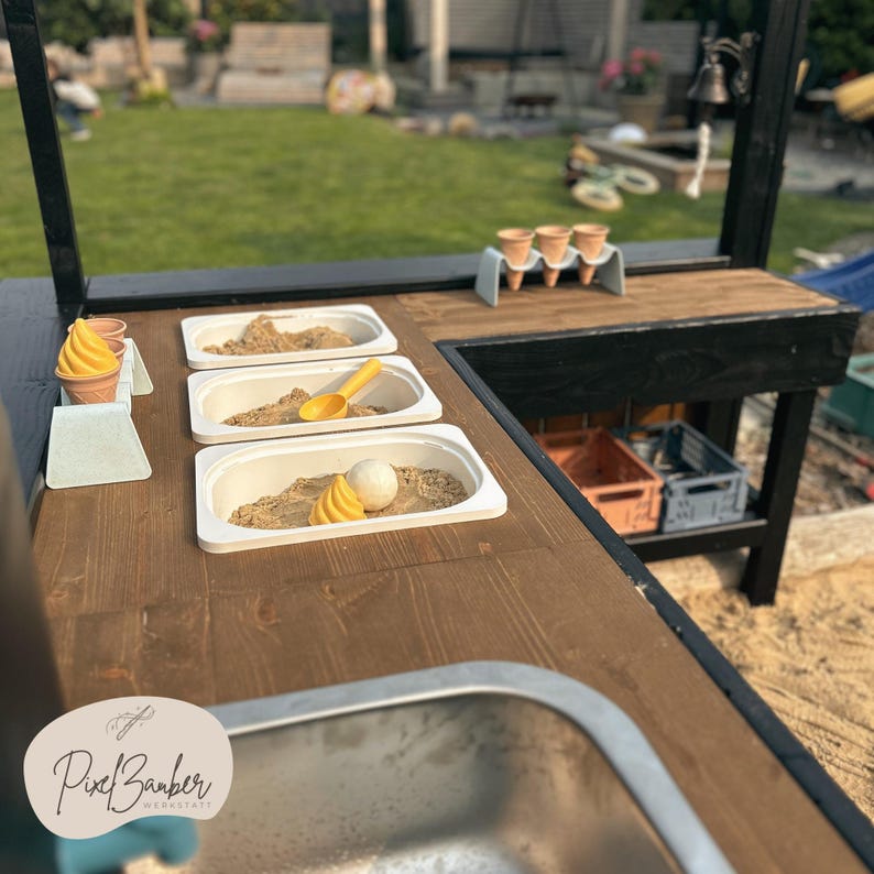 May include: A wooden play kitchen with a dark stained countertop and black frame. The kitchen features three white rectangular containers filled with sand, toy ice cream cones, and a yellow scoop. The PixelBarber logo is visible.