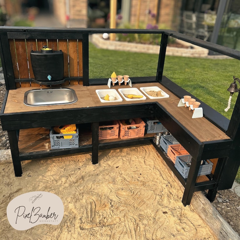 May include: A wooden outdoor play kitchen with a black frame and a natural wood countertop. It features a stainless steel sink, a water dispenser, and various containers with play food. The kitchen is set up in a sandbox.