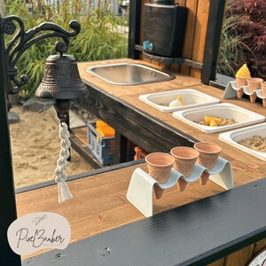 May include: An outdoor ice cream stand with a dark wooden counter and black trim. The stand features a metal bell, a sink, and containers with ice cream toppings. Empty ice cream cones are displayed on a white holder.