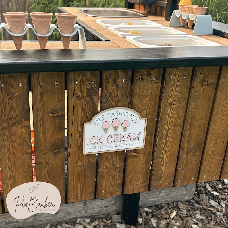 May include: Wooden ice cream stand with a sign that reads "Old Fashioned Ice Cream." The stand has a dark wooden top with containers for ice cream and a holder for ice cream cones. The sign also lists sprinkles, sundaes, and shakes.
