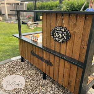 May include: A wooden outdoor stand with a black trim and legs. The stand has a sign that says "COME IN, WE'RE OPEN". The stand also has the word "Strandcafe" on the front. The stand is made of brown wood.