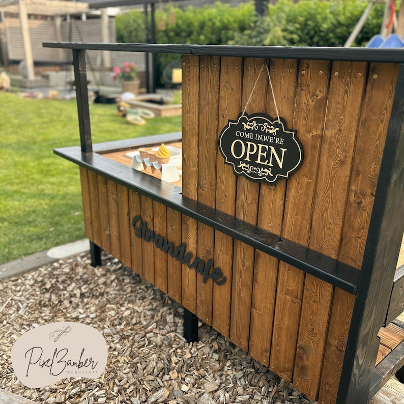 May include: A wooden outdoor stand with a black trim and legs. The stand has a sign that says "COME IN, WE'RE OPEN". The stand also has the word "Strandcafe" on the front. The stand is made of brown wood.