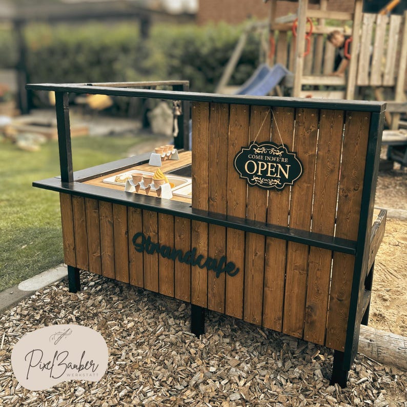 May include: A wooden ice cream stand with a black frame and the word "Strandcafe" in cursive. The stand has an "OPEN" sign and contains ice cream cones. The stand is outdoors on a bed of wood chips.