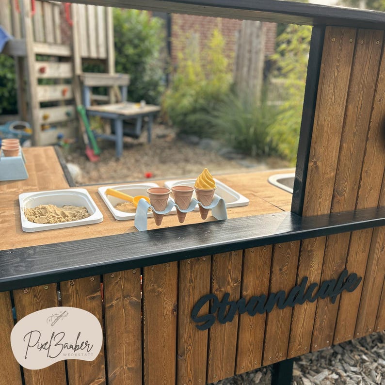 May include: A wooden play stand with a black trim and a sign that says "Strandcafe". The stand has a sand pit, a water basin, and three toy ice cream cones with a yellow swirl. The stand also has a sign that says "PixelBarber Werkstatt".
