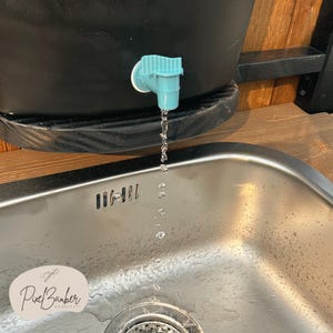 May include: A black water dispenser with a teal spigot, dispensing water into a stainless steel sink. The image includes the text "Pixel Barber Werkstatt".