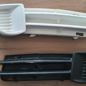 May include: Two car vent grilles, one white and one black, are displayed on a wooden surface. A digital caliper is positioned next to the white grille. The grilles have a slotted design and are likely automotive parts.