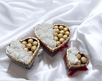 Pode incluir: Três caixas em forma de coração repletas de chocolates dourados e rosas de tecido branco. As caixas são adornadas com ouro e fita vermelha, sobre um fundo de cetim branco. Uma ideia de presente romântica.