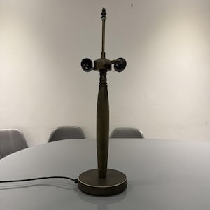 Może przedstawiać: Brązowa lampa stołowa z wąską, toczoną drewnianą podstawą i okrągłą podstawą. Lampa ma dwa gniazda żarówek i ozdobny szczyt na górze. Lampa jest podłączona do gniazdka elektrycznego.