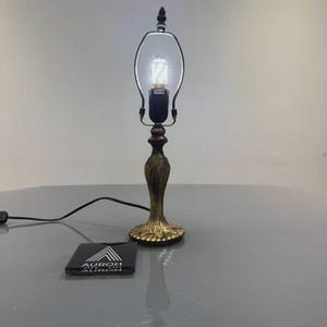 Op de afbeelding: Een decoratieve messingkleurige tafellamp met een heldere glazen kap en een zwart snoer. De lamp heeft een sierlijke voet en een kleine, decoratieve bekroning aan de bovenkant. Een klein zwart vierkant met de tekst "AUROH" staat aan de voet van de lamp.
