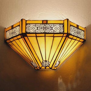 May include: A stained glass wall sconce in a geometric shape. The sconce has a warm yellow and orange colour scheme, with black lines and decorative patterns. A decorative lighting fixture.