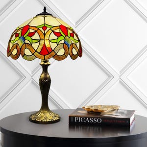 Może przedstawiać: Lampa stołowa z witrażowym kloszem w kształcie kopuły, ozdobionym czerwonymi, zielonymi i kremowymi szklanymi panelami w geometryczny wzór. Lampa posiada podstawę z ciemnego metalu w kolorze brązu z dekoracyjnym, fakturowanym trzonem i stopą.