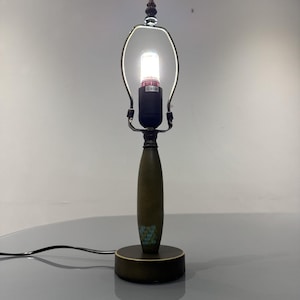May include: A vintage table lamp with a dark green base and stem, decorated with small turquoise accents. The lamp has a metal shade holder and a bright, illuminated bulb. It is connected to a black power cord.