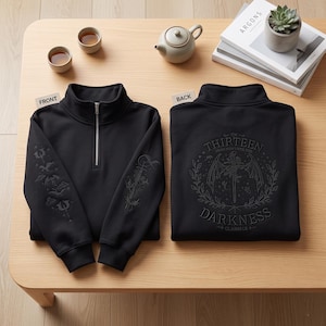Peut inclure: Un sweat-shirt noir à fermeture éclair au quart avec des détails brodés sur les manches et un grand motif au dos. Le dos présente une épée, des ailes et des fleurs avec le texte "THE THIRTEEN DARKNESS CLAIMS US" disposé en cercle.
