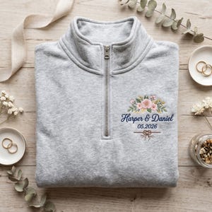 May include: A light gray quarter-zip sweatshirt with embroidered floral design and the names "Harper & Daniel" with the date "05.2026". The sweatshirt is folded and placed on a wooden surface.