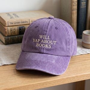 May include: A purple baseball cap with a distressed, washed-out finish. The front of the cap features the embroidered text "WILL YAP ABOUT BOOKS" in a cream-coloured, serif font. The cap has a curved brim and a structured six-panel crown design.