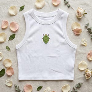 May include: A white ribbed sleeveless crop top featuring a small, centered embroidery of a green leaf-shaped insect with brown legs and antennae. The garment has a high, rounded neckline and a fitted silhouette, showcasing a clean, simple design.