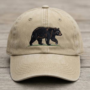 May include: A tan baseball cap featuring an embroidered design of a black bear walking on a thin green line. The cap has a structured crown, a curved brim, and stitched eyelets on the upper panels, providing a classic look and functional design.
