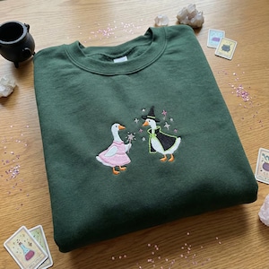 May include: Dark green sweatshirt featuring embroidered geese. One goose is in a pink dress and holds a wand, the other wears a witch hat and cape. The design includes stars. A comfortable, whimsical garment. Made from soft fabric.