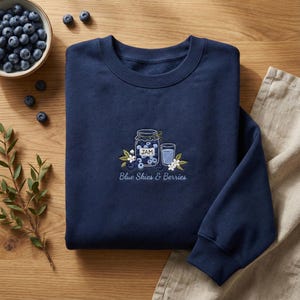 May include: A navy blue sweatshirt featuring an embroidered design of a jam jar, a glass, blueberries, and flowers. The text "Blue Skies & Berries" is below the design. A bowl of blueberries and greenery are in the background.