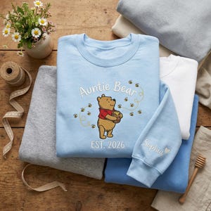 May include: A light blue sweatshirt featuring a Winnie the Pooh design with the text "Auntie Bear" and "EST. 2026". The sleeve has the name "Sophia" embroidered. Other sweatshirts in white and gray are stacked nearby.