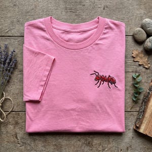 Peut inclure: T-shirt rose à col rond et manches courtes. Un motif de fourmi rouge et noire est brodé sur le devant. Le t-shirt est plié sur une surface en bois avec des éléments décoratifs.