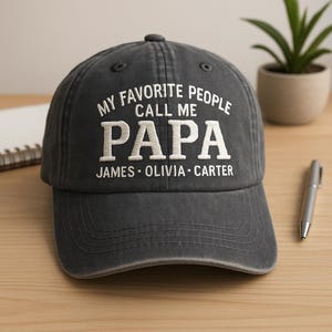 May include: A charcoal gray baseball cap with the embroidered text "MY FAVORITE PEOPLE CALL ME PAPA" in white. Below the word "PAPA" are the names "JAMES • OLIVIA • CARTER". The cap has a curved brim and a fabric strap.