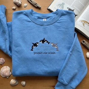 Könnte beinhalten: Hellblaues Sweatshirt mit gestickten Orcas und dem Text "protect our ocean". Das Design zeigt drei Orcas, zwei in Schwarz-Weiß und einen in Grau, mit stilisierten Wellen. Ein lässiges Kleidungsstück zum Thema Ozean.